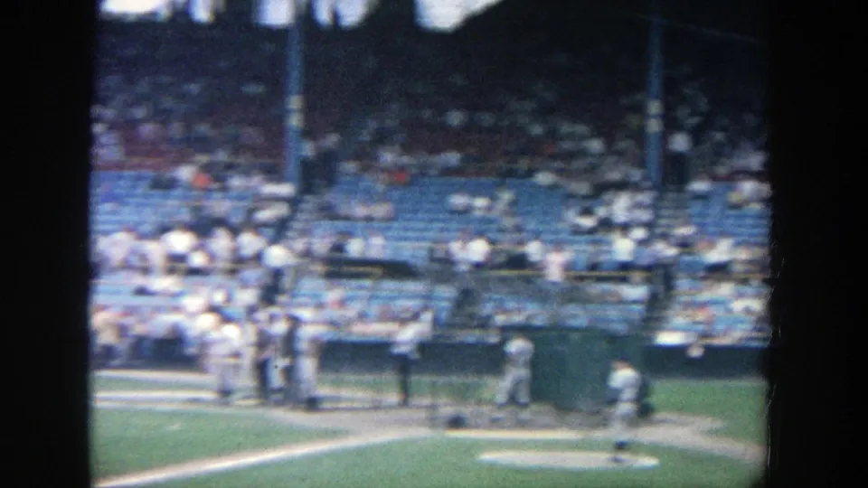 Home-movie frame from 1964 Yankee Stadium showing players around the infield edge and spectators seated behind the field.