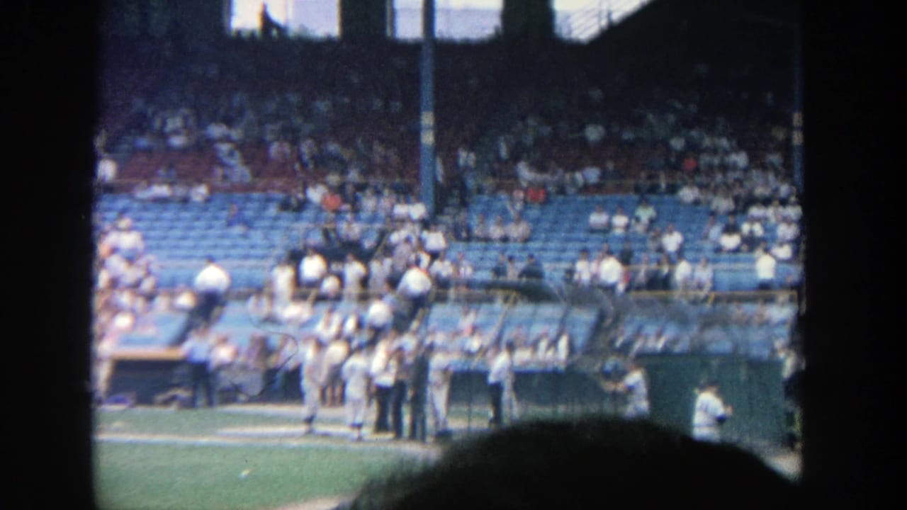 1964 Yankee Stadium home-movie frame showing fans in blue seating and uniformed players near the field-level dugout area.