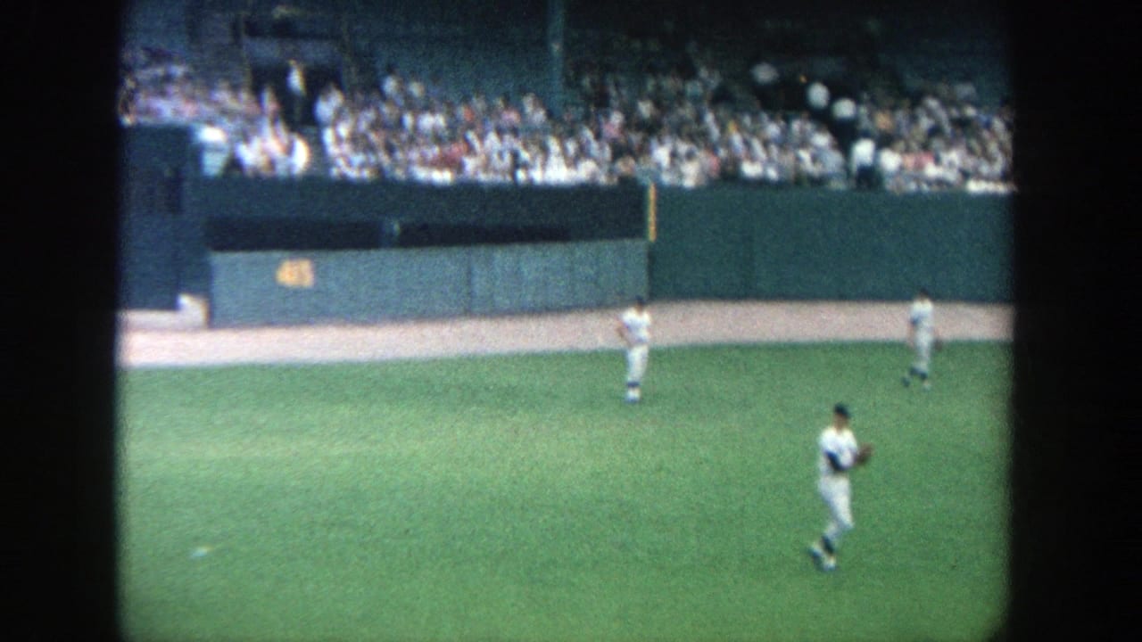 1964 Yankee Stadium home-movie frame showing outfielders below crowded seating and a visible yellow distance marker on the wall.