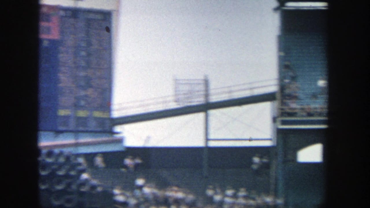 1964 Yankee Stadium home-movie frame showing part of the scoreboard, commercial signage, railings, and spectators in the stands.