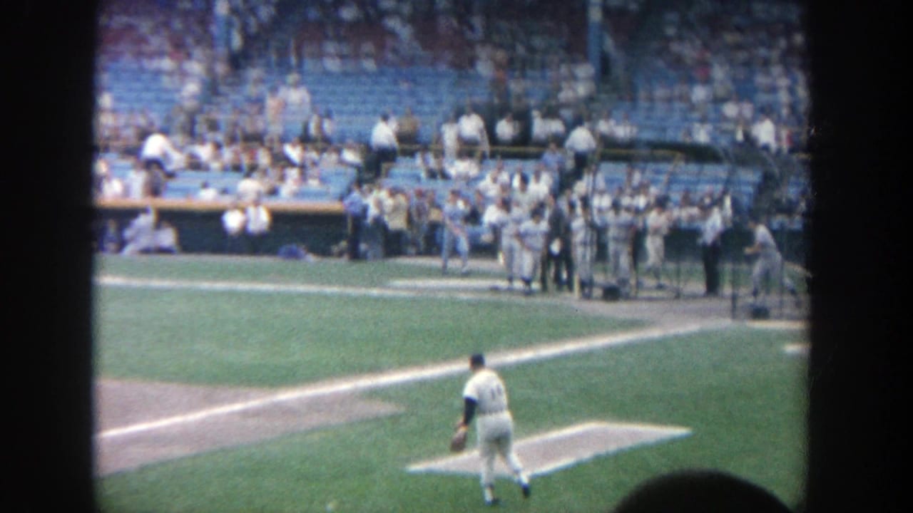 1964 Yankee Stadium home-movie frame showing a pitcher near the mound, dugout activity, and spectators in blue seats.