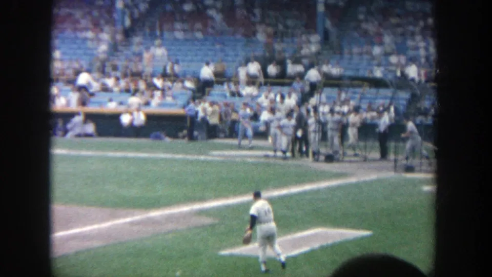 1964 Yankee Stadium home-movie frame showing a pitcher near the mound, dugout activity, and spectators in blue seats.