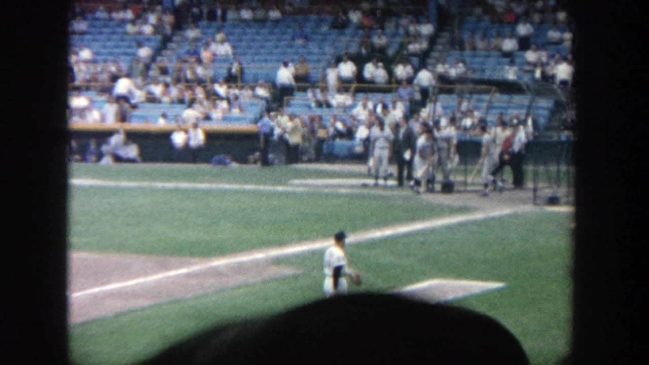 Baseball field view with a pitcher, dugout area, and spectators seated behind the field.