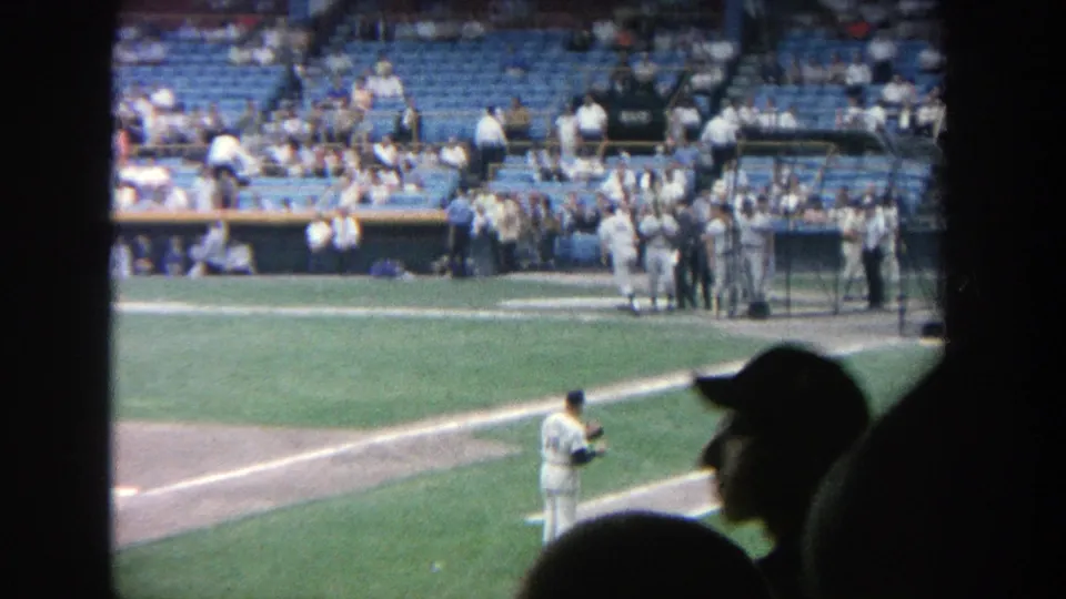 Spectator-shot Yankee Stadium frame with a pitcher in white uniform, dugout activity, and audience silhouettes in the foreground.