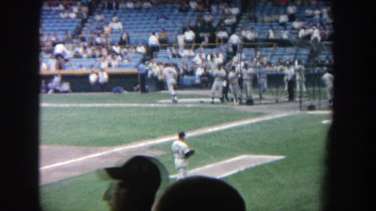 Baseball field seen from the stands with players near home plate and spectators in blue seats.