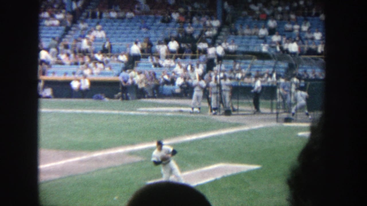 Pitcher or fielder in a white uniform stands on the grass with home plate area behind him.