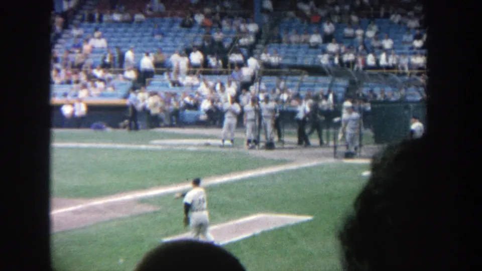 Spectator-shot 1964 ballpark frame showing a player from behind, the home plate area, dugout activity, and blue seating at Yankee Stadium.