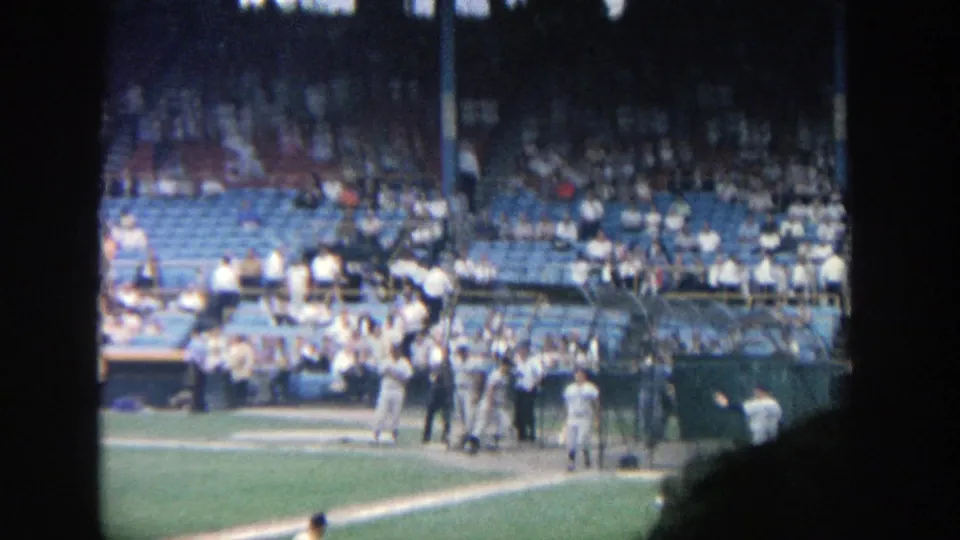 Wide 8mm frame of Yankee Stadium showing a batter near home plate, nearby players, dugout area, and spectators in the blue seats.