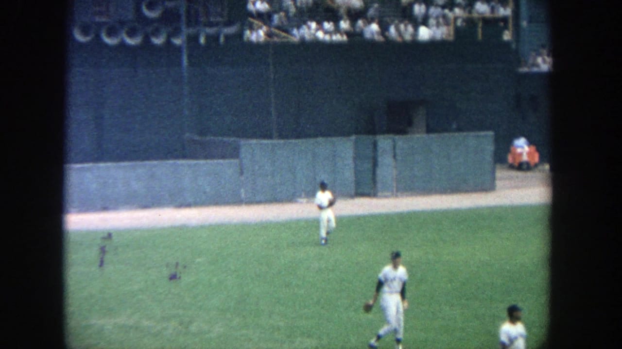 1964 Yankee Stadium frame with players positioned across the grass in front of the outfield wall and distant spectators.