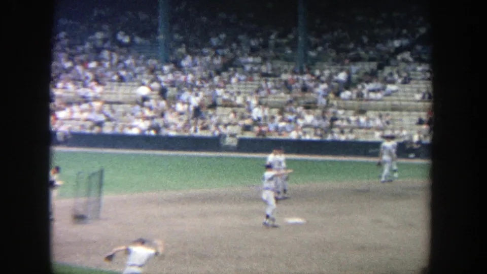 1964 8mm view of baseball players warming up on the Yankee Stadium infield, with a crowded seating section in the background.