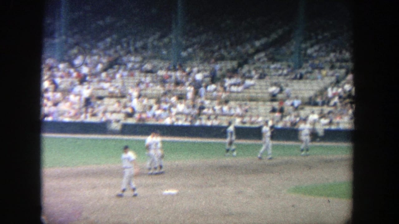 Uniformed players stand and move near the infield with crowded stadium seating behind them.