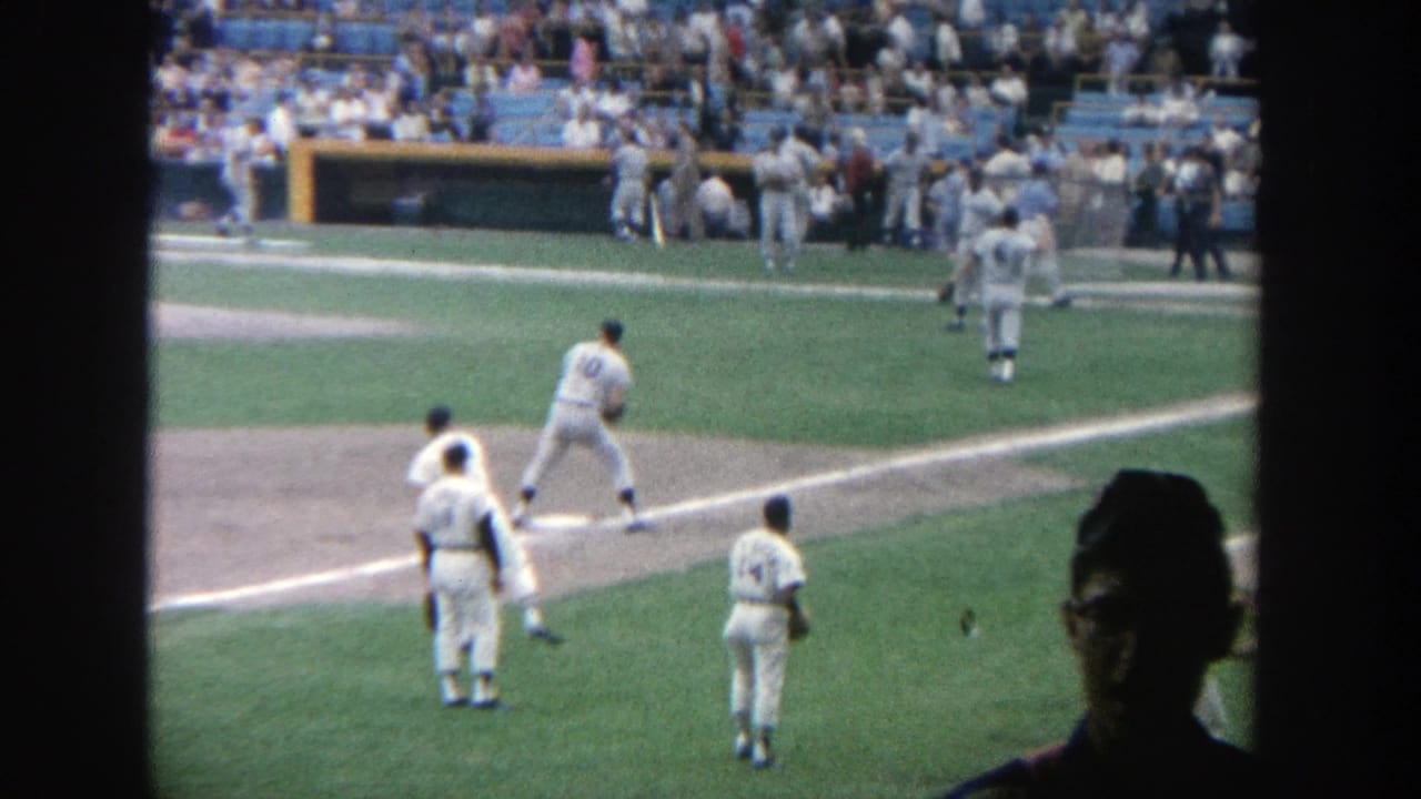 1964 Yankee Stadium home-movie frame showing players near the infield, blue seating, spectators, and a dark foreground silhouette.