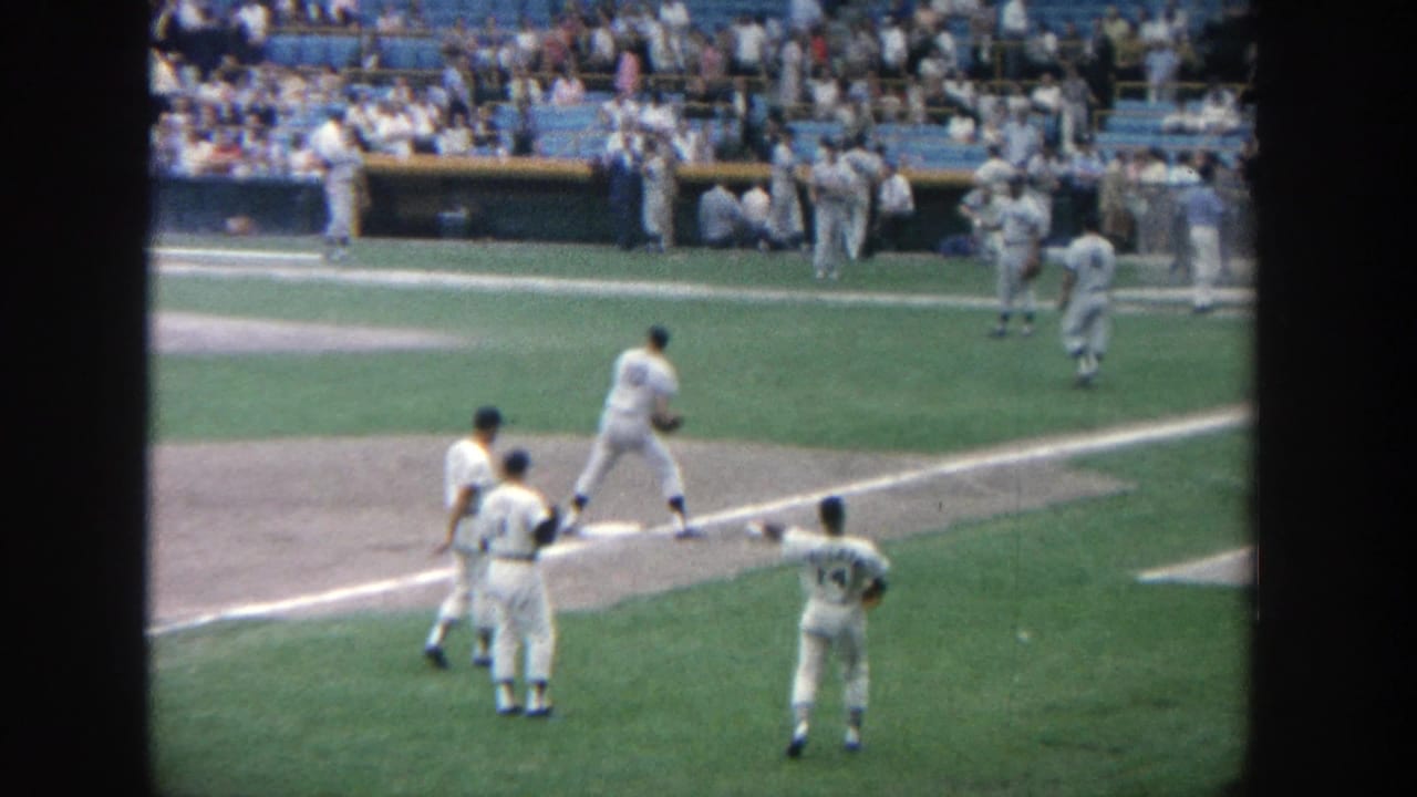 Players in white uniforms move near the base path while spectators fill blue seats in the background.
