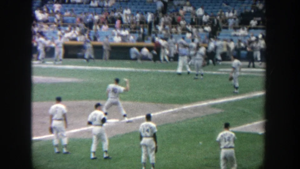 1964 Yankee Stadium home-movie view of several players in white uniforms near the infield with spectators and dugout activity behind them.
