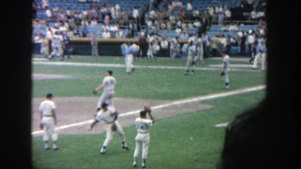 1964 Yankee Stadium frame showing players around the infield, a dark foreground edge, dugout activity, and spectators in blue seats.