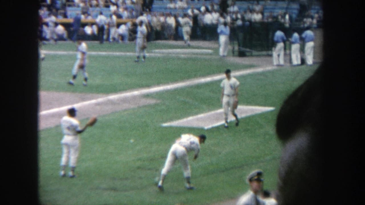 1964 Yankee Stadium frame with baseball players warming up, a dugout area, spectators, and a dark foreground edge.