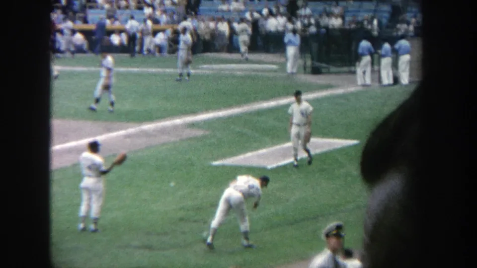 1964 Yankee Stadium frame with baseball players warming up, a dugout area, spectators, and a dark foreground edge.