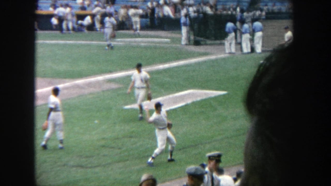 1964 Yankee Stadium frame showing a baseball player mid-throw during warmups, with teammates, spectators, and field lines visible.