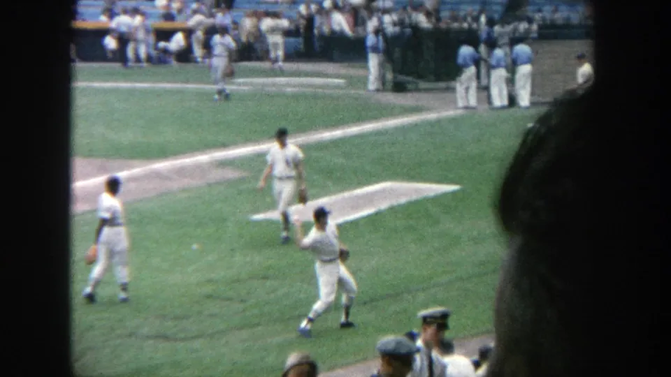 1964 Yankee Stadium frame showing a baseball player mid-throw during warmups, with teammates, spectators, and field lines visible.