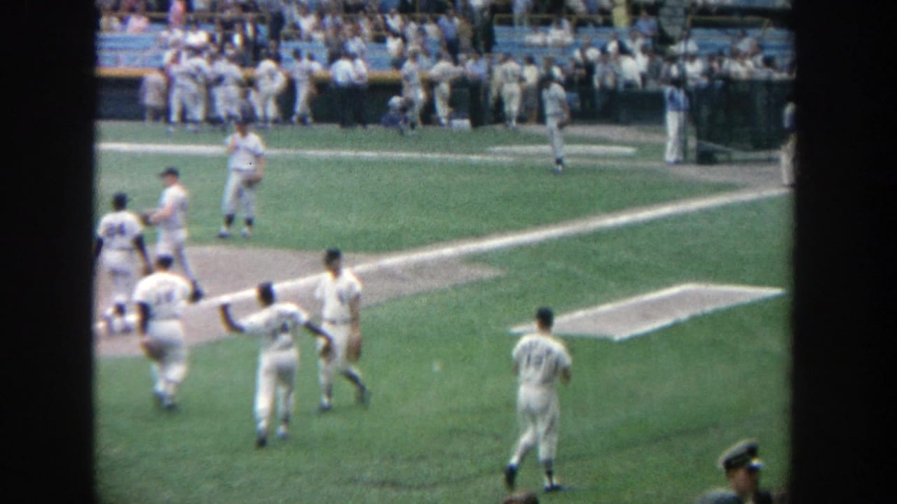 Spectator-shot 1964 Yankee Stadium frame showing several baseball players near the infield and filled stands in the background.