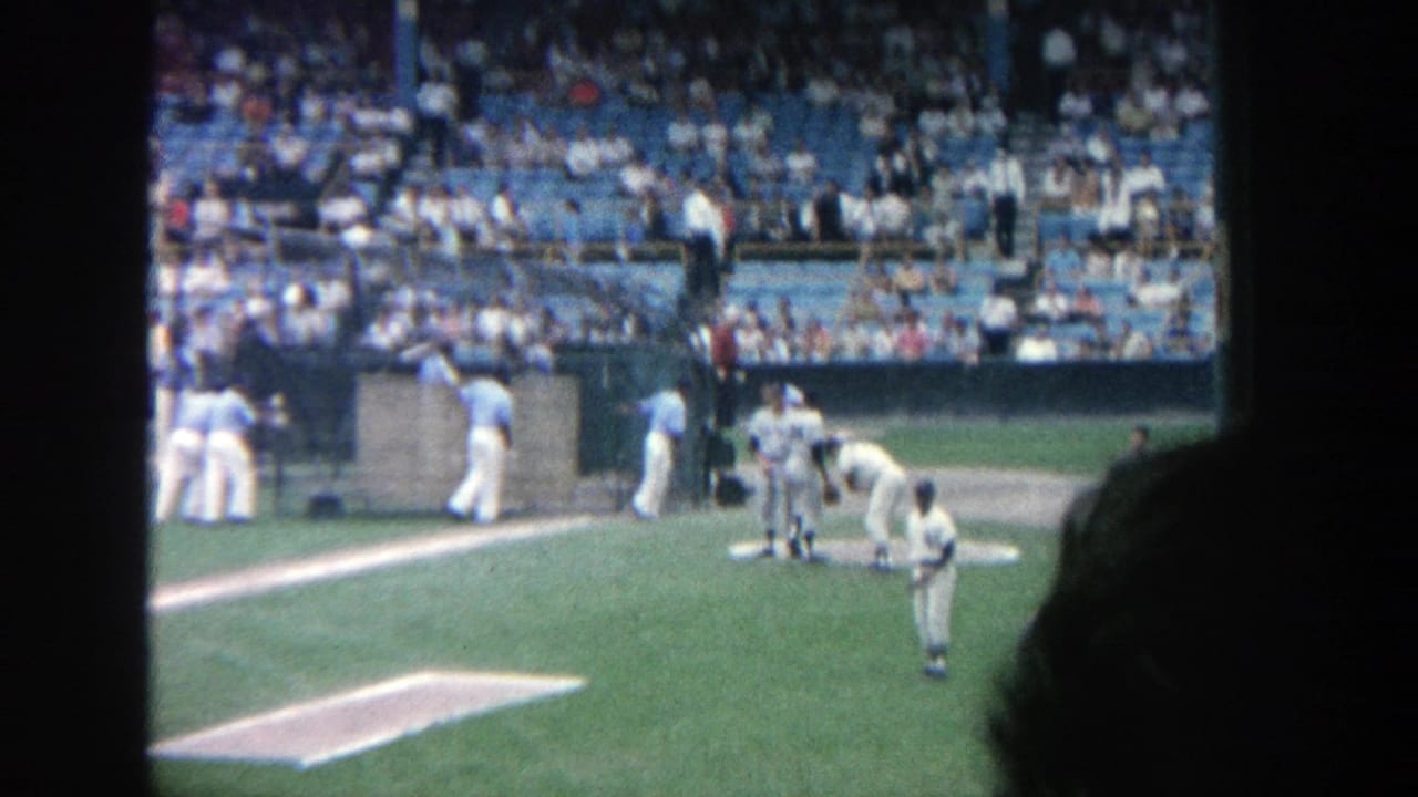Wider 1964 Yankee Stadium view showing players near the mound and infield, with blue seats and spectators beyond.