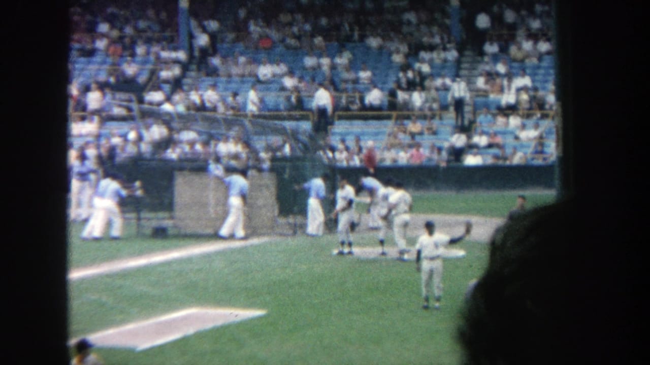 1964 8mm view of Yankee Stadium with players near a dugout or field structure and a crowd watching from blue seats.
