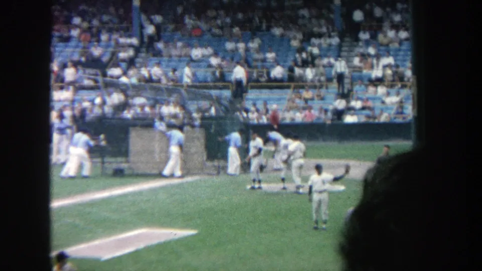 1964 8mm view of Yankee Stadium with players near a dugout or field structure and a crowd watching from blue seats.