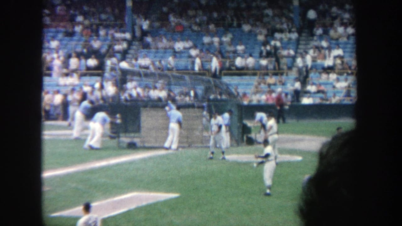 Players in light uniforms stand near a field structure with spectators in the stands behind them.