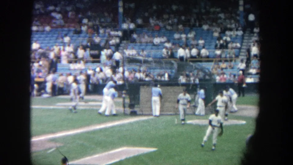 Spectator-shot 8mm frame at Yankee Stadium with a foreground player and others clustered near the dugout-side structure.