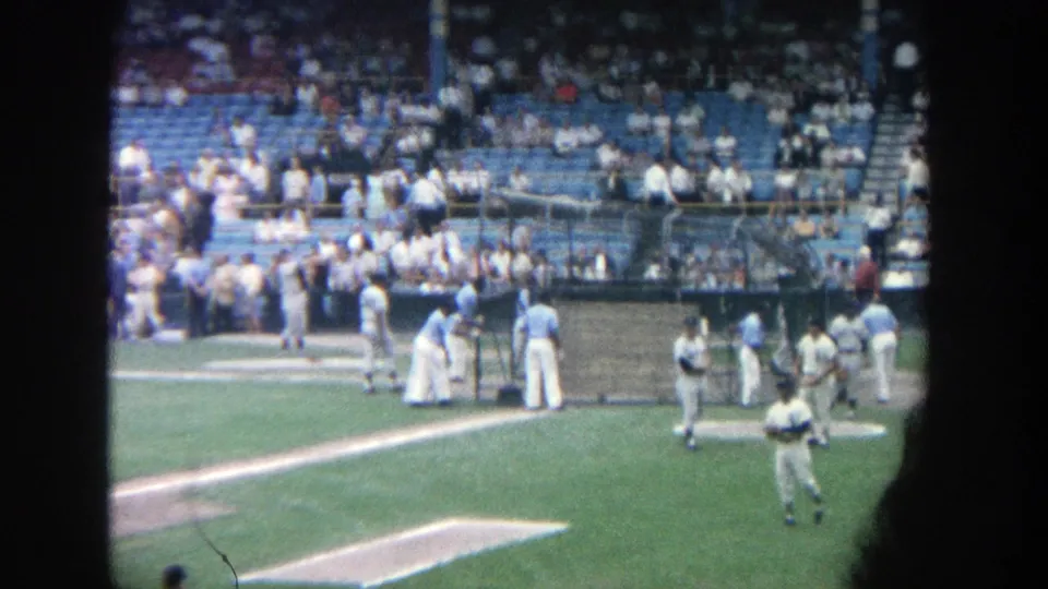 1964 Yankee Stadium home-movie frame with players near the dugout area and spectators seated behind them.