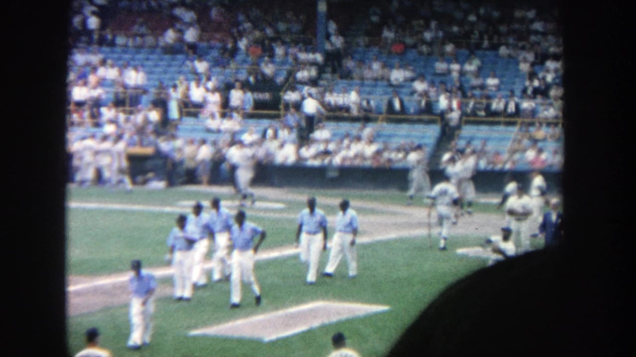 Baseball players in light uniforms walk near the infield with blue stadium seats behind them.