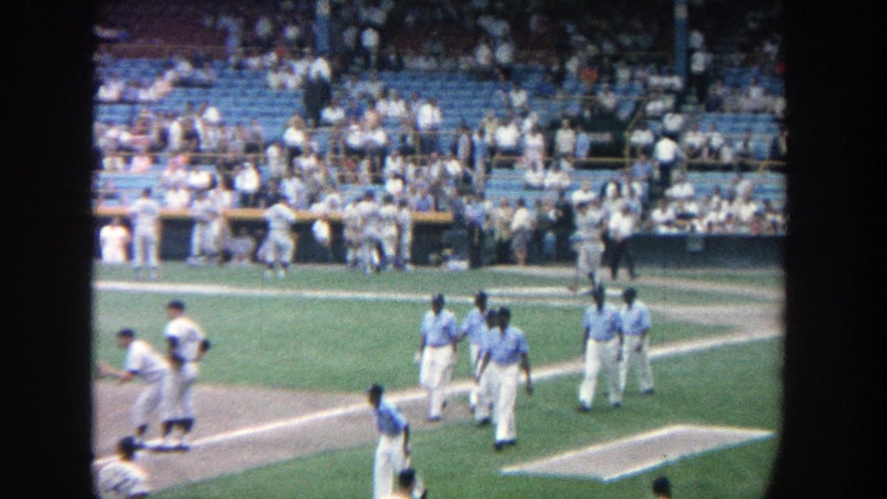 Uniformed baseball players move near home plate and the dugout with spectators in the stands.