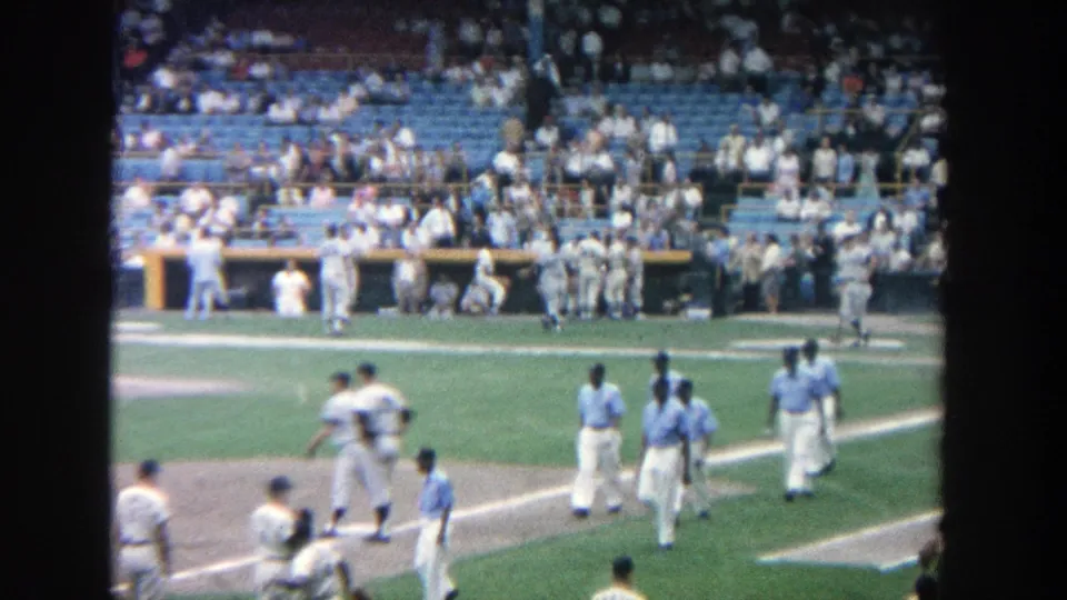 The 1964 home-movie frame shows players in light uniforms walking near the infield edge at Yankee Stadium, with the dugout and crowd behind.