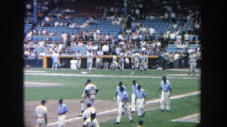 At Yankee Stadium in 1964, several uniformed players cross the field near the infield while spectators sit behind the dugout area.