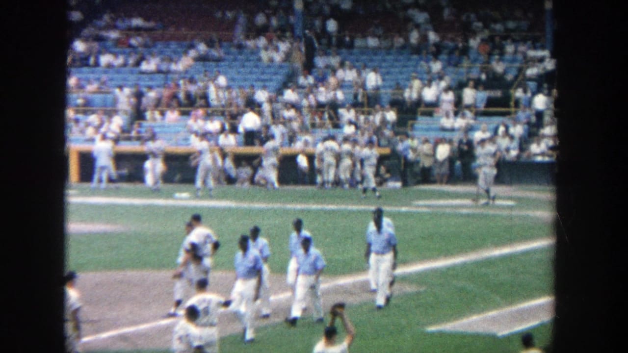1964 Yankee Stadium home-movie frame showing players near the infield and dugout area as spectators watch from blue seats.
