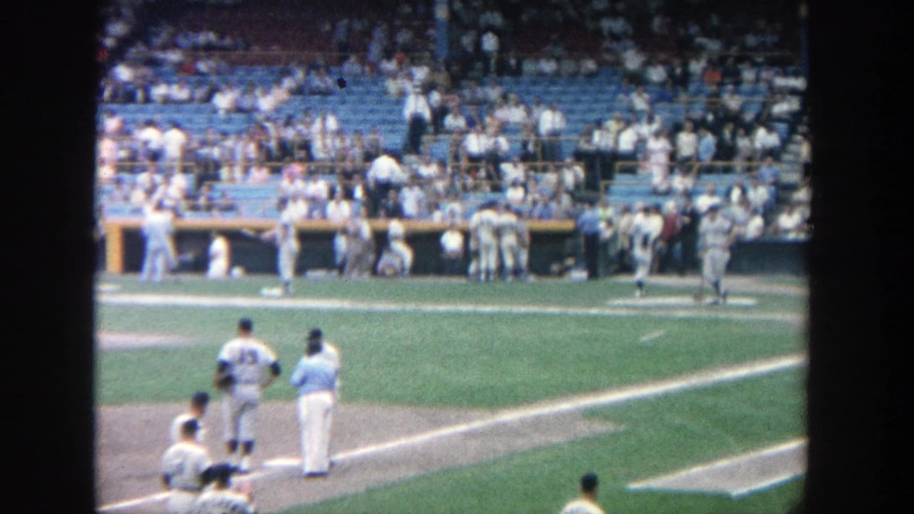 1964 Yankee Stadium frame with players near the first-base side and a busy dugout and seating area behind them.