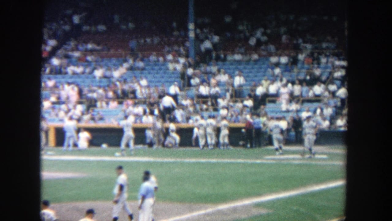 1964 Yankee Stadium home-movie frame showing a player moving across the field with dugout activity and spectators behind him.