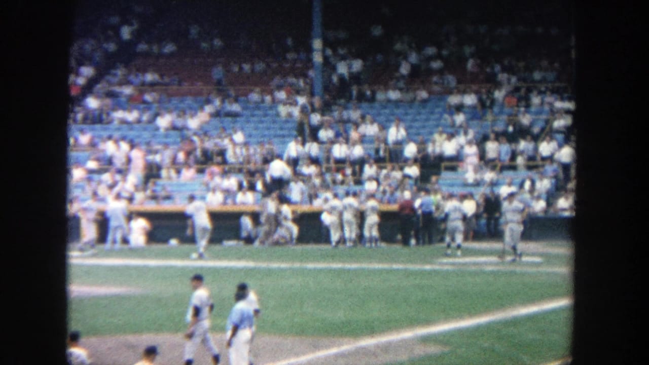 1964 home-movie view at Yankee Stadium showing players near the foul line, dugout activity, and spectators in blue seats.