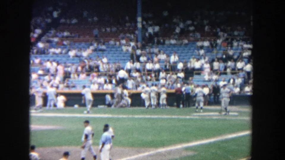 1964 home-movie view at Yankee Stadium showing players near the foul line, dugout activity, and spectators in blue seats.