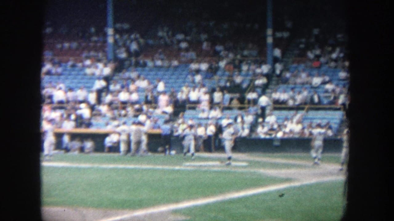 Players and spectators are visible near the dugout and first-base side of a baseball field.