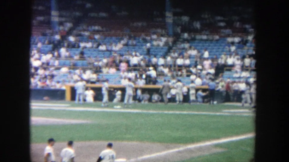 1964 Yankee Stadium frame showing players near the infield dirt, dugout area, blue seats, and a scattered crowd.