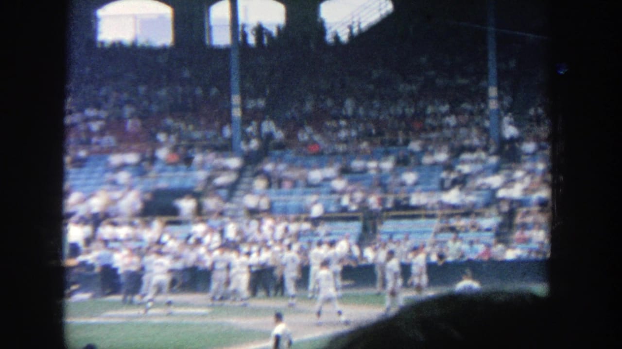 Baseball players in white uniforms gather near the dugout with spectators in the stands.
