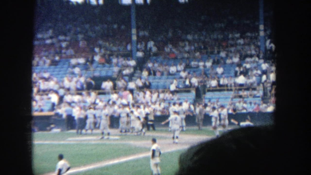 Players stand and walk near the dugout while a large crowd watches from the stands.