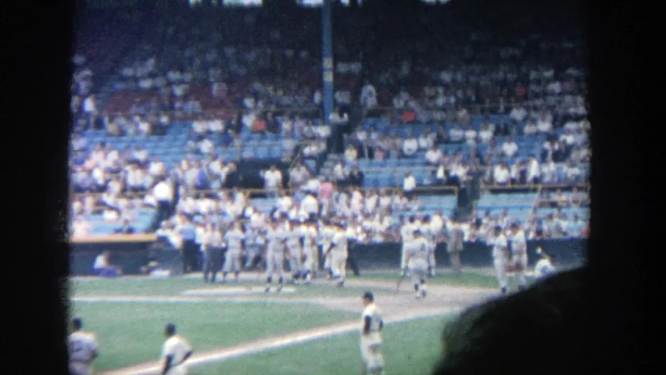 1964 Yankee Stadium home-movie still showing a group of white-uniformed players by the dugout and a full section of spectators.