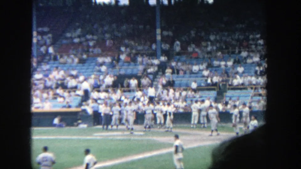1964 Yankee Stadium frame with players near the dugout, a crowded grandstand, and dark home-movie frame edges.