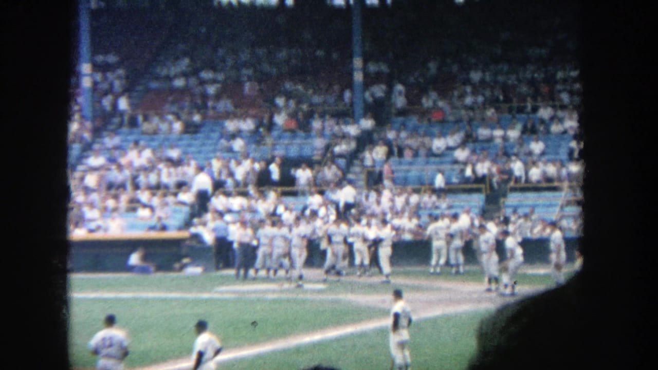 1964 Yankee Stadium home-movie frame showing baseball players near the infield and a crowd in the stands.