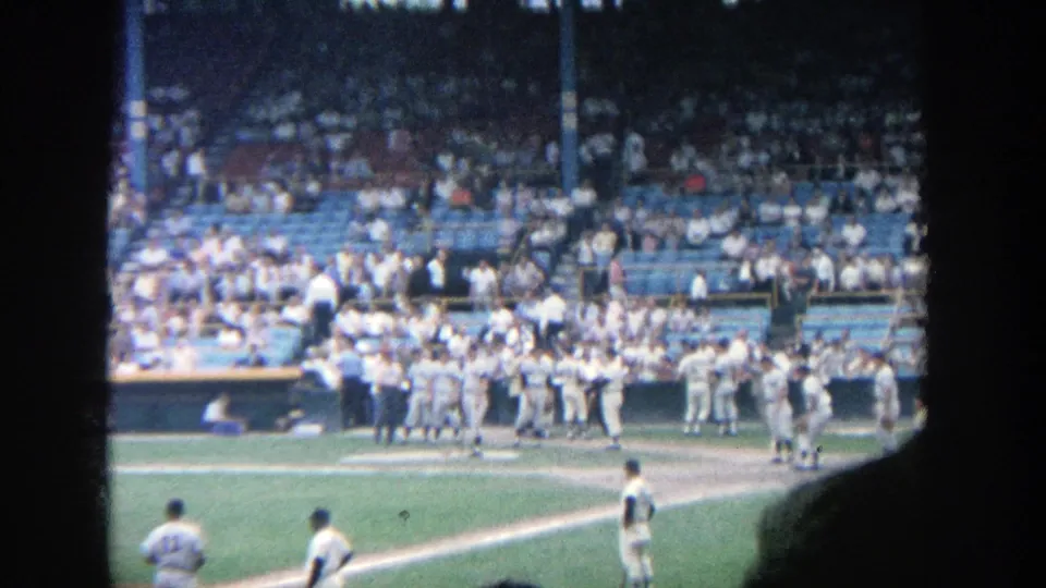 1964 Yankee Stadium home-movie frame showing baseball players near the infield and a crowd in the stands.