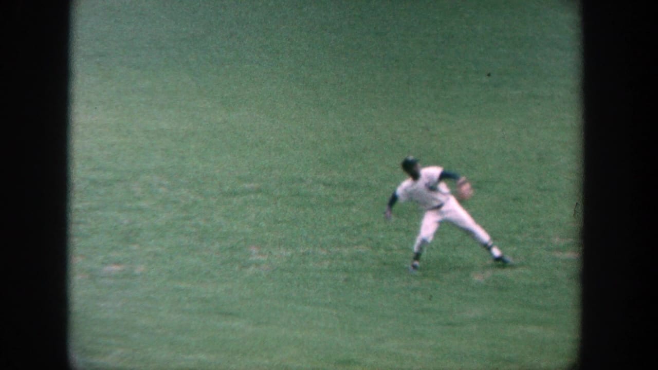 A baseball player in a white uniform throws or fields on the grass outfield.