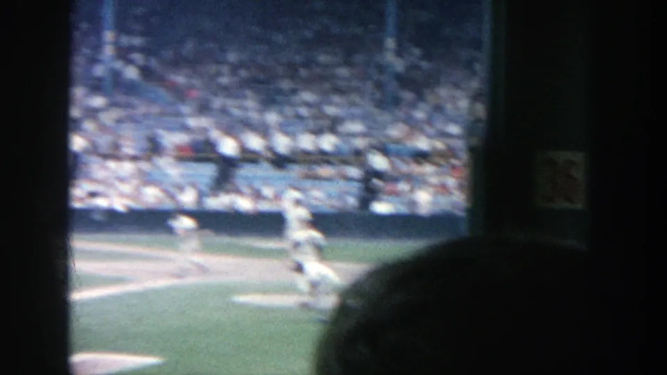 A handheld 8mm view shows baseball players near the infield with stadium spectators visible in the background.