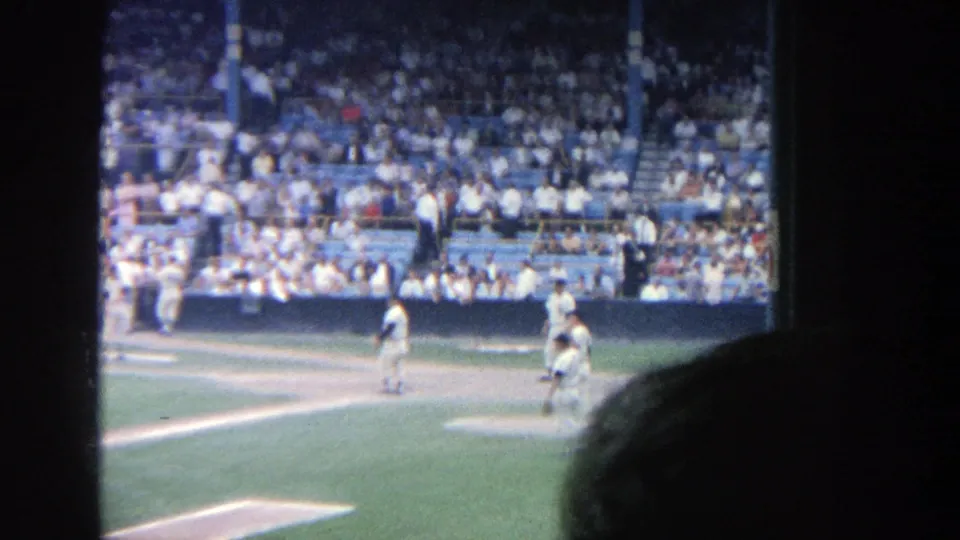 Players stand near the infield at Yankee Stadium, with a seated crowd visible in the stands in a 1964 8mm frame.